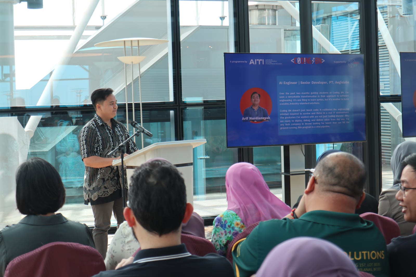 The author at a podium giving a speech, with a slide on the screen behind him showing his bio: AI Engineer and Senior Developer at PT. Aegislabs, next to the Coding BN logo.