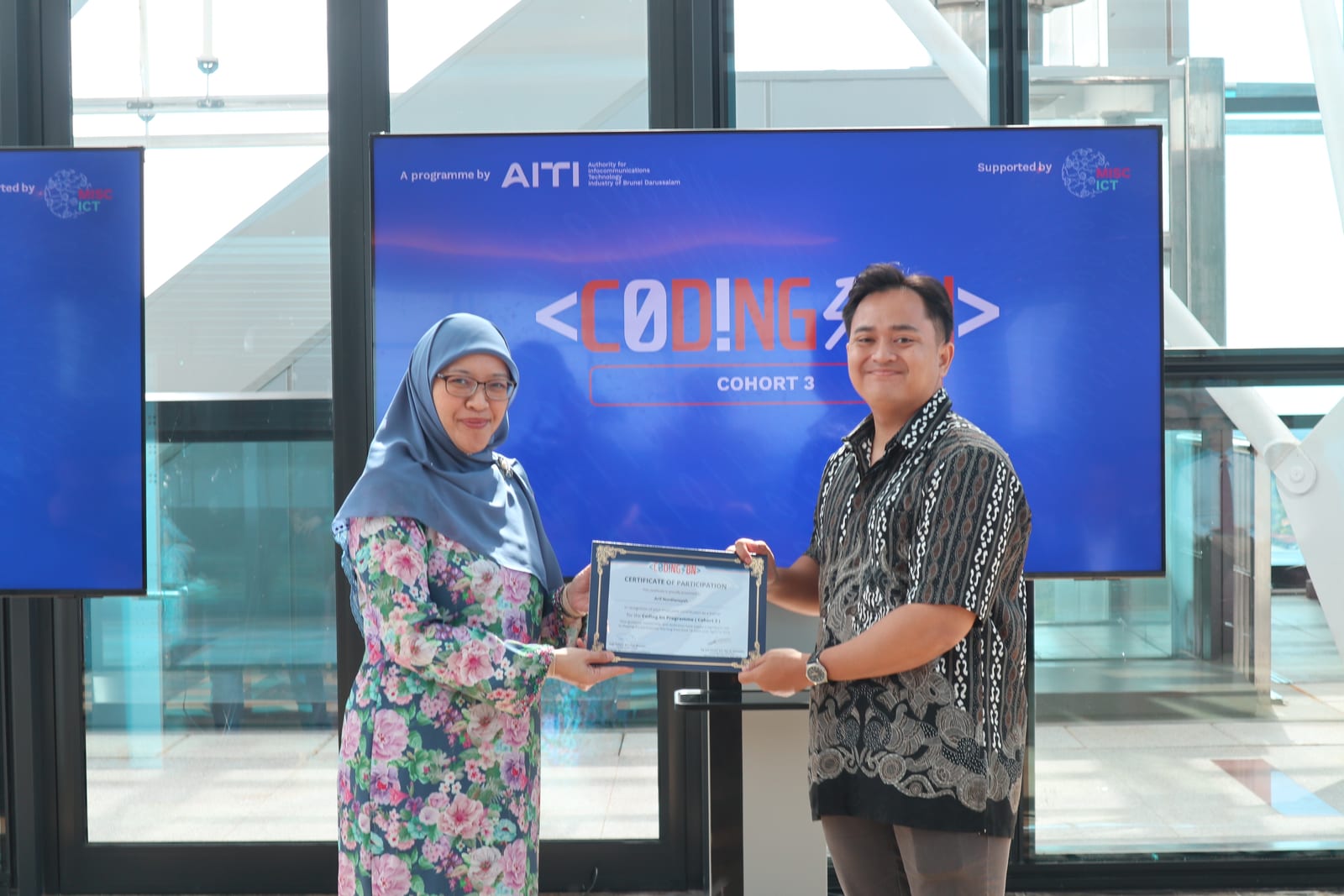 The author receiving a certificate of recognition from a Ministry official in formal Brunei attire, in front of a backdrop showing the Coding BN Cohort 3 branding.